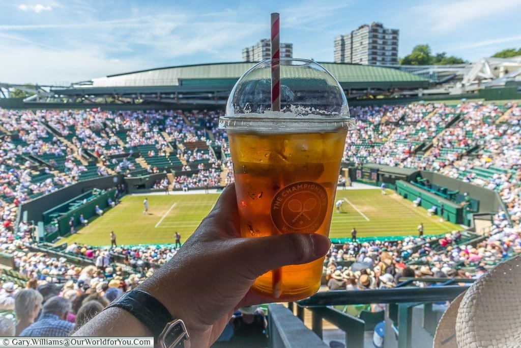 A clear plastic beaker with domed lid and straw, full of Pimms and lemonade, held aloft in front of a game of tennis at the centre court of wimbledon during the finals.