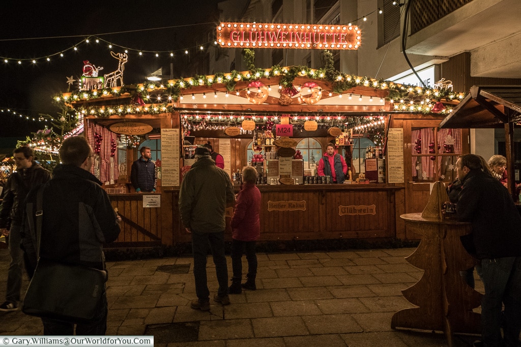 A brightly lit Glühwein hut in Frankfurt's quayside Christmas market at night.  People are coming and going, others are perched around wooden tables, shaped like Christmas trees enjoying their drinks.
