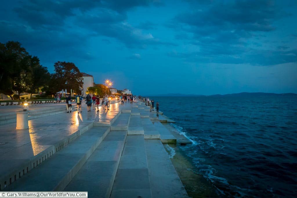 The Sea Organ, hidden under Zadar’s Riva at dusk, after the sun has set, but there is still blue light.  Shallow steps lead down to the waters of the Adriatic Sea lapping against the edge.