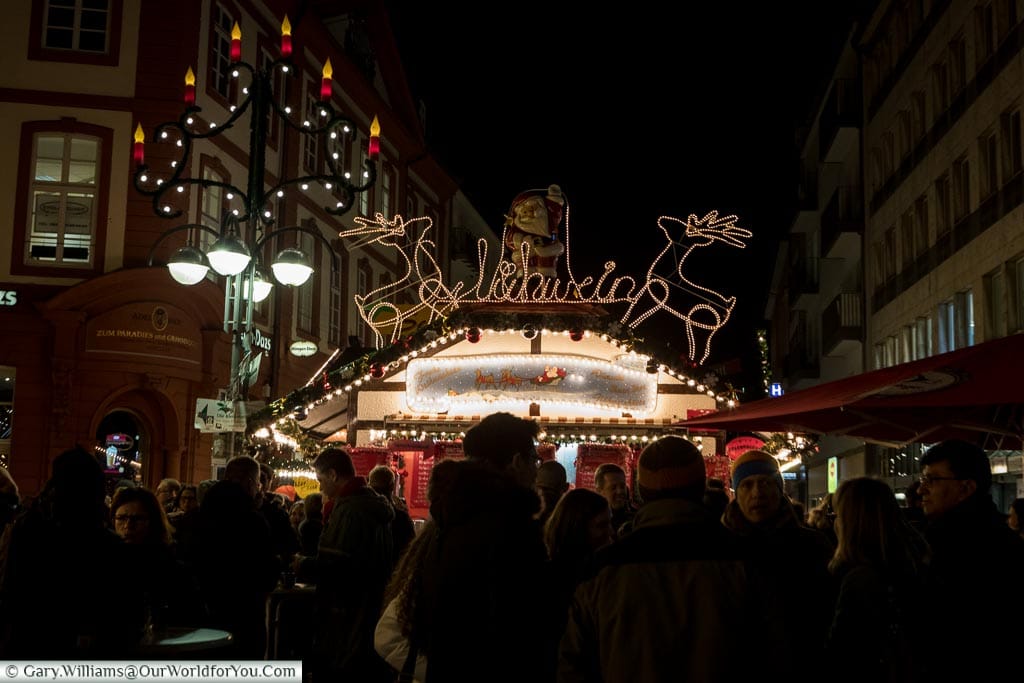 A group of people gathered around a glühwein stall on the Christmas market at night.  The streel lamp has an additional advent candlelight attached, and the top of the booth has two illuminated reindeer either side of Santa and a glühwein sign.