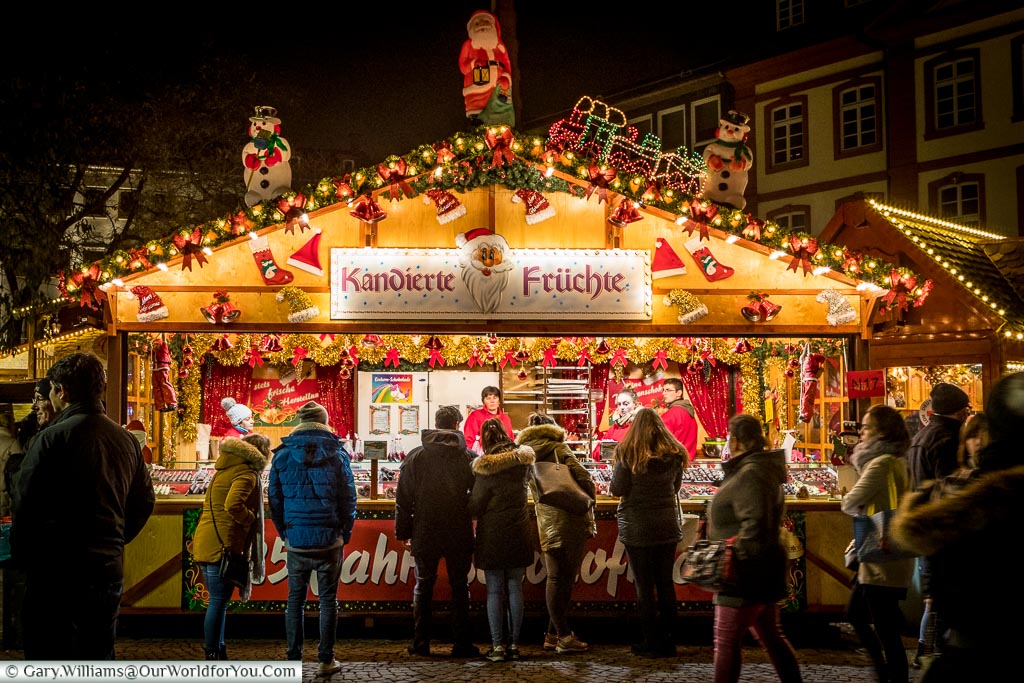 A brightly lit, candied fruit stall, on Frankfurt’s Christmas market. People gather to look at the selection of wares.