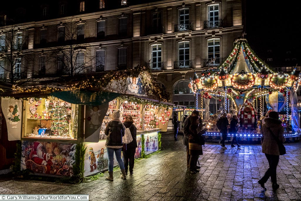 A small group of people gather between a Christmas market stall and the carousel in Place De La Cathédrale.