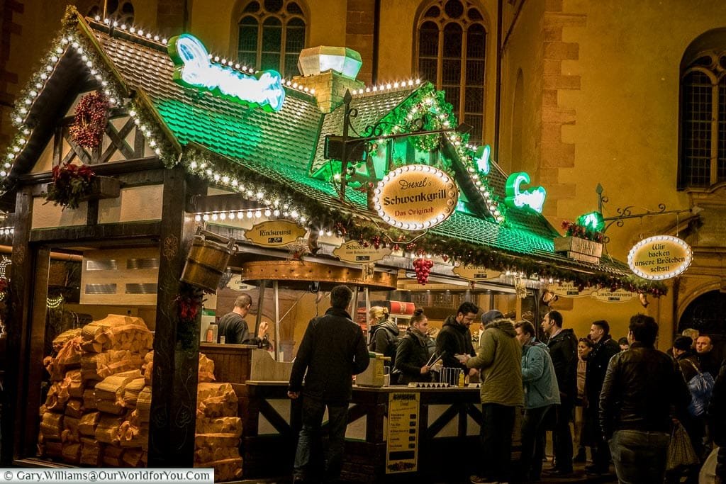 A food stall at night at the Christmas market in front of St. Catherine's Church. The food is cooked over an open wood-fired grill and you can see the sacks of logs stacked at the end of the cabin.