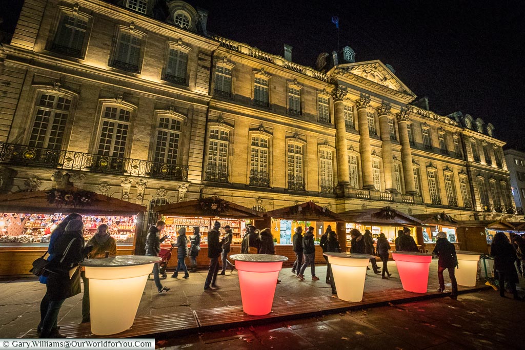 People gathering around illuminated tables that resemble giant plastic beakers in front of the Christmas market 'Marche-aux-Poissons' in front of the floodlit Palais Rohan.