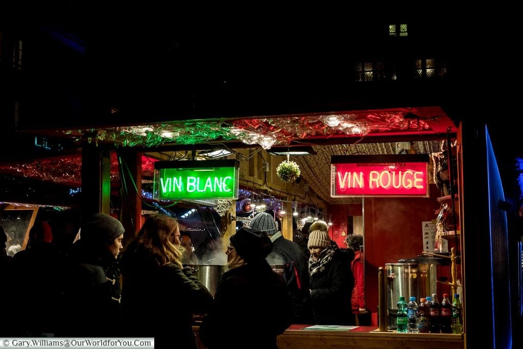 At the edge of a Christmas market drinks stall in the Place De La Cathédrale. There are two neon signs, one offering vin blanc, the other vin rouge.