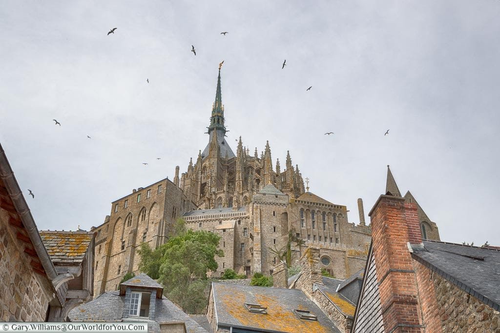 Birds circle the spire of the Mont-Saint-Michel Abbey, high on the hill, as the skies darken.