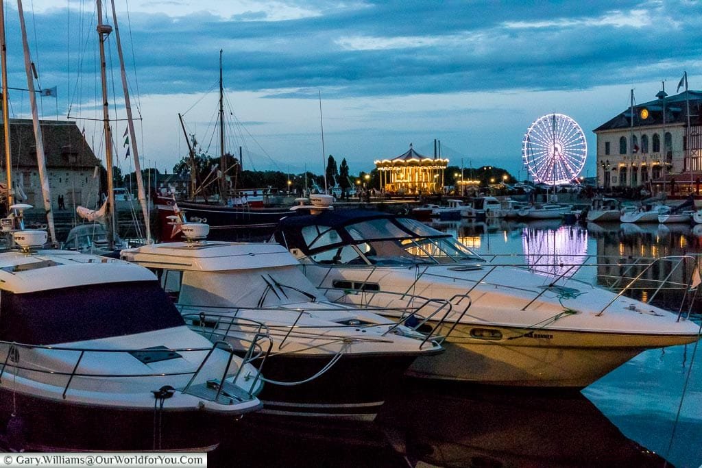 Boats in Honfleur's harbour at dusk with the illuminated carousel and Ferris wheel in the background.