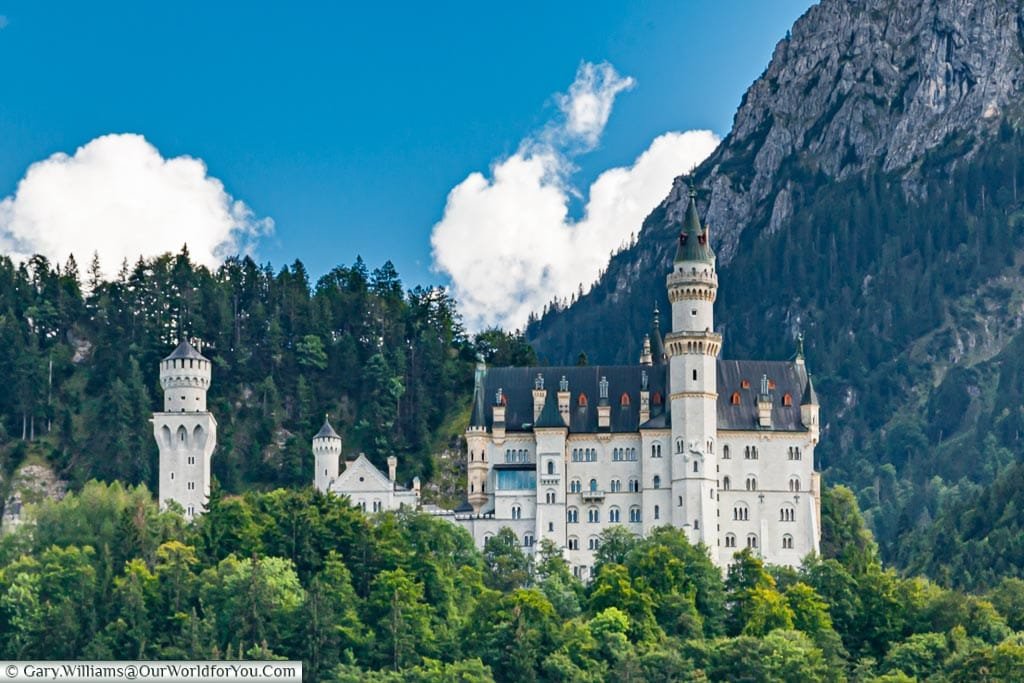 A side view of white stone Neuschwanstein Castle set amongst the forests and the mountains of southern Bavaria.  This fairy tale castle was the inspiration for Walt Disney’s Magic Kingdom castle.