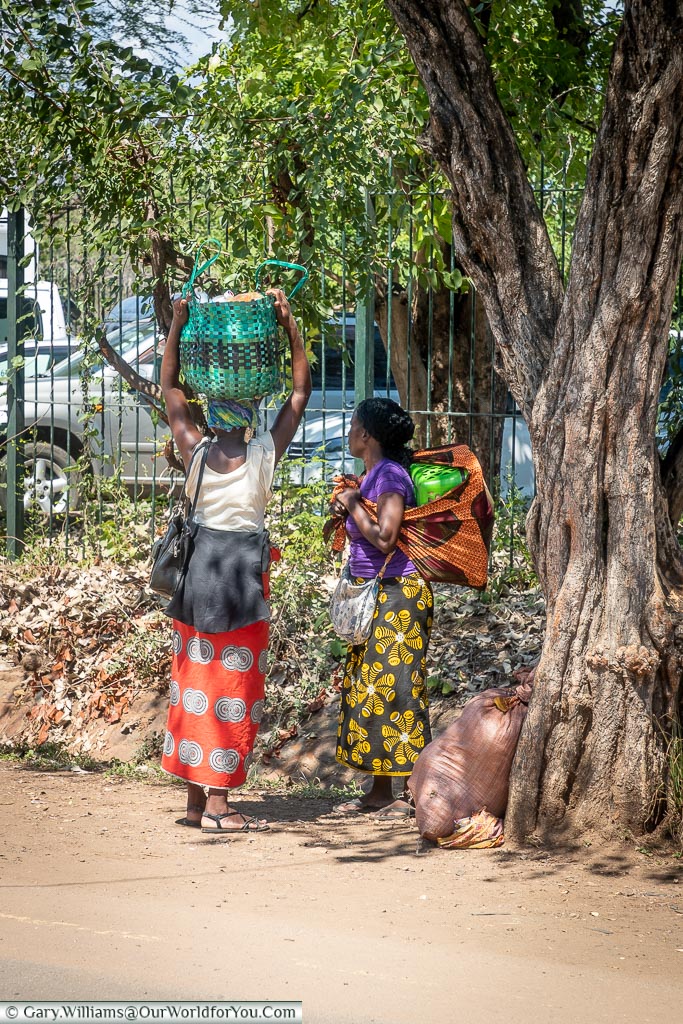 Two women carrying heavy loads, with one balancing it on her head, in the town at Victoria Falls, Zimbabwe