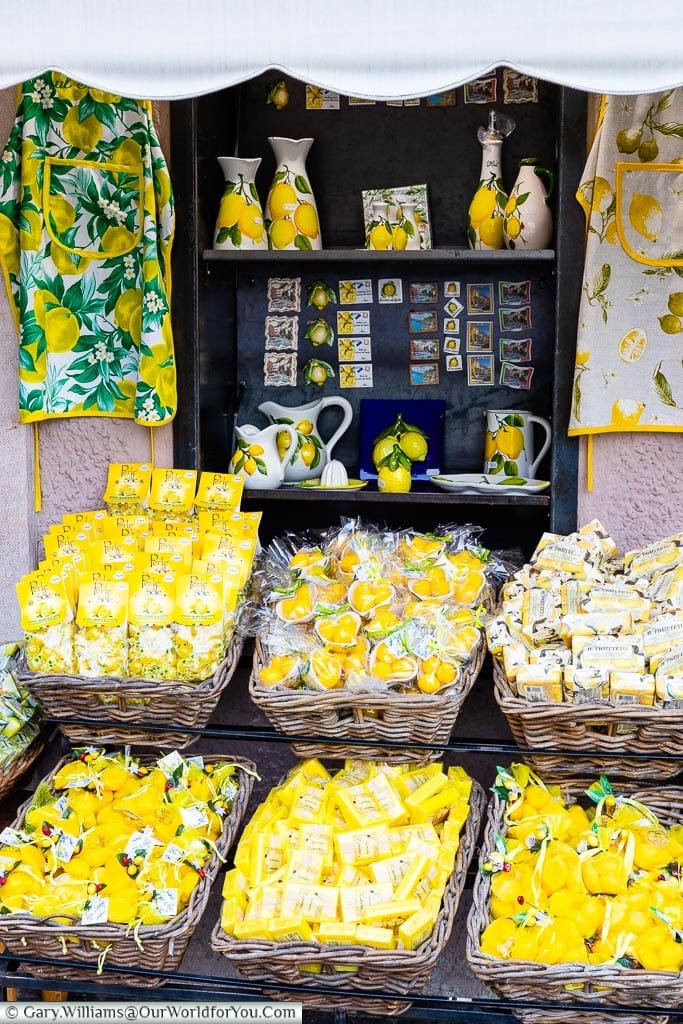 The display outside a shop with baskets of Lemon soaps, bath bombs, lemon sweets & other lemon based products. On the shelves behind are jugs, olive oil poured, fridge magnets all decorated with lemons.