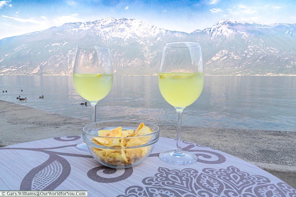 Two glasses of Limoncello Sprits, with plain crisps, next to the edge of Lake Garda with the mountains as a backdrop.