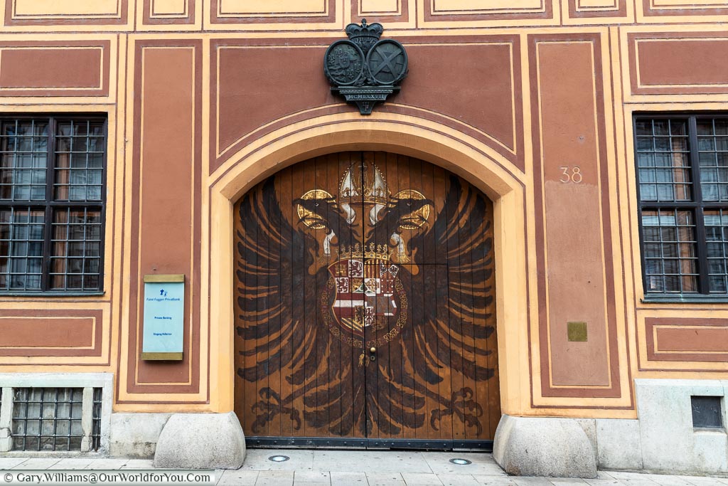 A pair of large wooden gates, decorated with a twin-headed eagle and a heraldic shield that mark the entrance to the Fürst Fugger Privatbank.