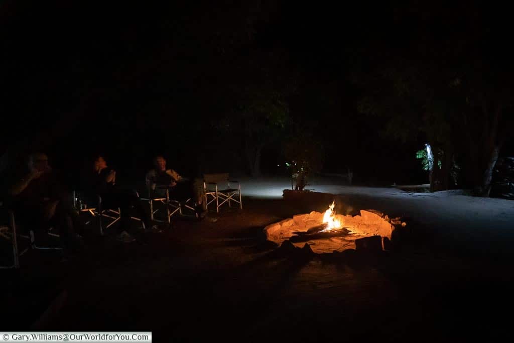 A small group sitting around an open-air fire pit, with a roaring fire.