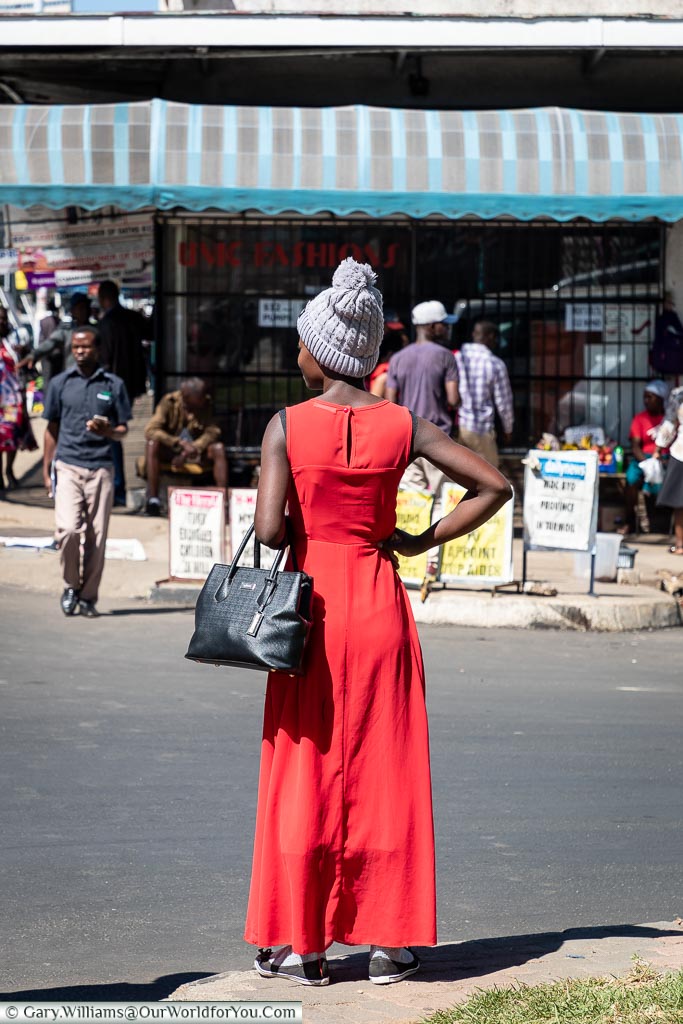 A woman in a full length, sleeveless, red dress wearing a grey wool bobble hat on a warm day in central Harare.