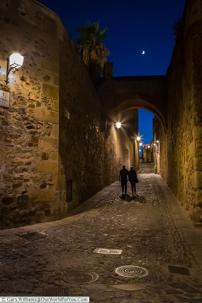 A couple walking a quiet lane in Cáceres lit by lanterns .