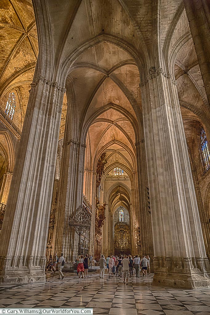 A portrait view inside Seville's gothic cathedral with a group at the base of one of its vast columns that support its substantial vaulted ceiling.