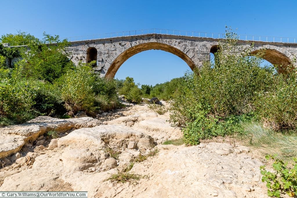 The small Roman bridge over a dry riverbed against a clear blue sky.