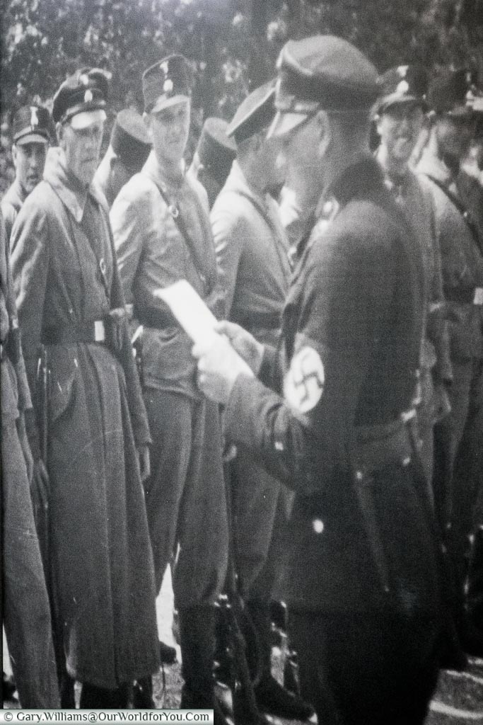 A blown-up black & white image of an officer wearing a swastika armband addressing a group camp guards.