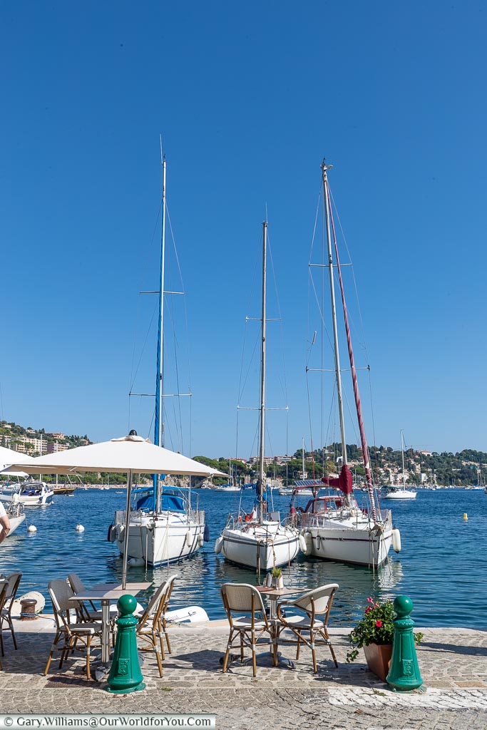 A table & chairs at the edge of the harbour of Villefranche-sur-Mer in front of 3 small yachts.