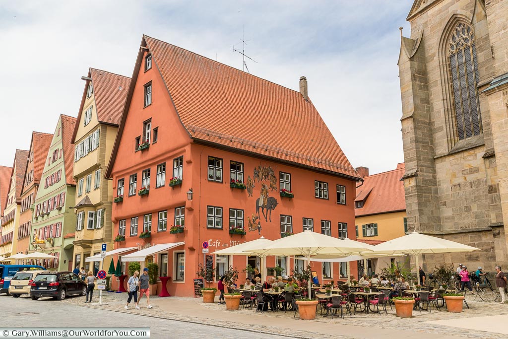 Tables and chairs under parasols in front of the colourful Café am Münster.  The picturesque buildings are painted in traditional colours, the cafe supporting a historic looking mural.