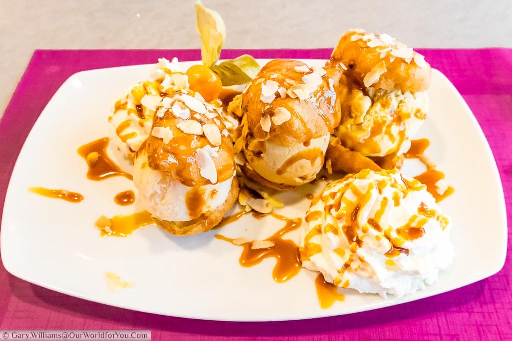 Three profiteroles stuffed with different flavoured ice creams topped with toasted almonds with  a dollop of whipped cream on the side, all drizzled with caramel sauce.
