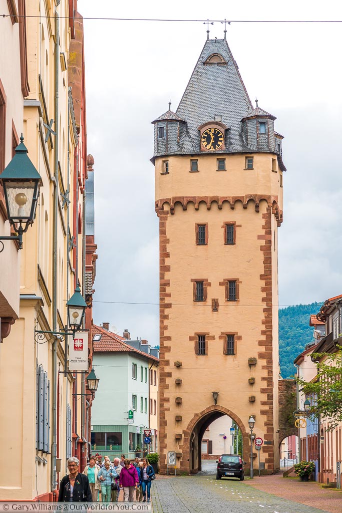 A tall gothic gatehouse at one end of the high street in Miltenberg.  The orange brick tower is topped with a grey tiled roof incorporating look out points and a clock.