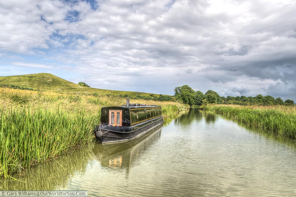 A black narrowboat mode up against the reeds on the Kennet and Avon canal in Wiltshire on a beautiful idyllic sunny day under a light cloudy sky