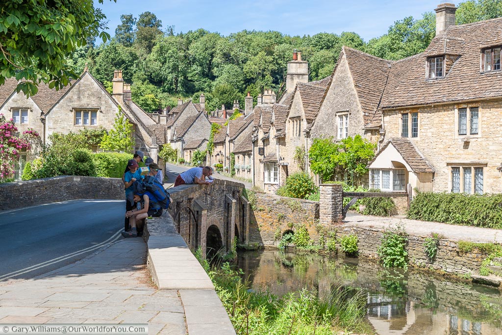 A family gathered on a small stone bridge over the By Brook in Castle Combe.  The road leading over the bridge he is flanked on either side my beautiful stone buildings in the traditional Cotswold style of a golden sandy stone.