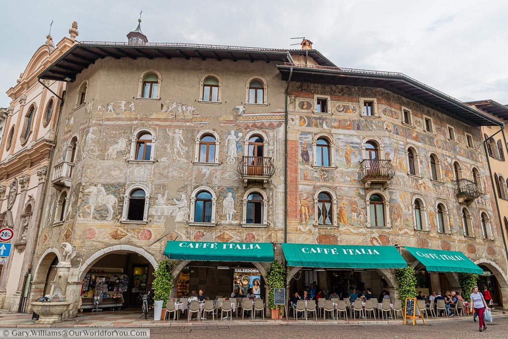 Café Italia in Piazza Duomo in the centre of Trento with green awnings covering the outdoor seating area with beautifully painted walls depicting classical Italian figures on the three storey renaissance building