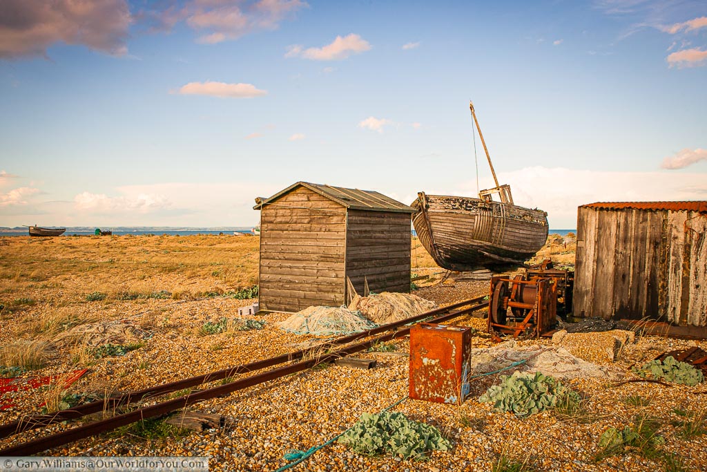 A scene of abandoned fishermans’ equipment including a wooden trawler, wooden shacks, rron winches and small track railway lines on the beach of Dungeness on the Kent coastline in England