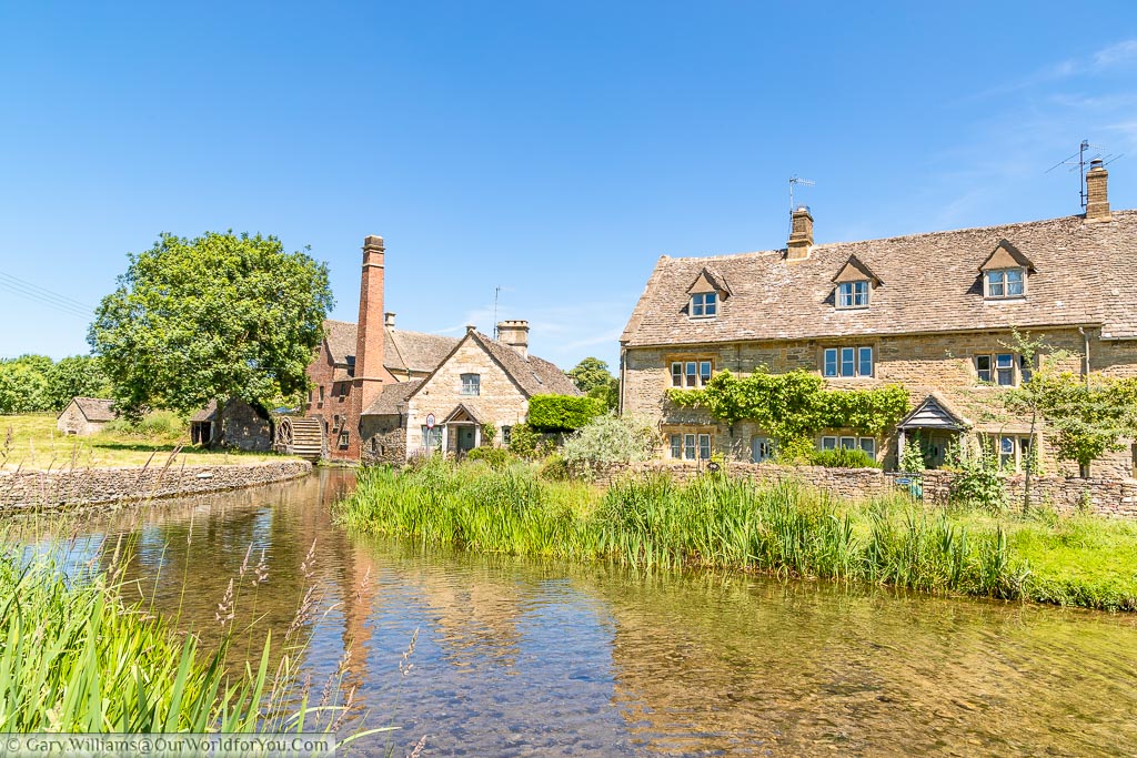 An idyllic Cotswold scene of a very shallow, slow flowing, river in front of a Cotswold home and a water mill with a tall brick built chimney In a village called Lower Slaughter