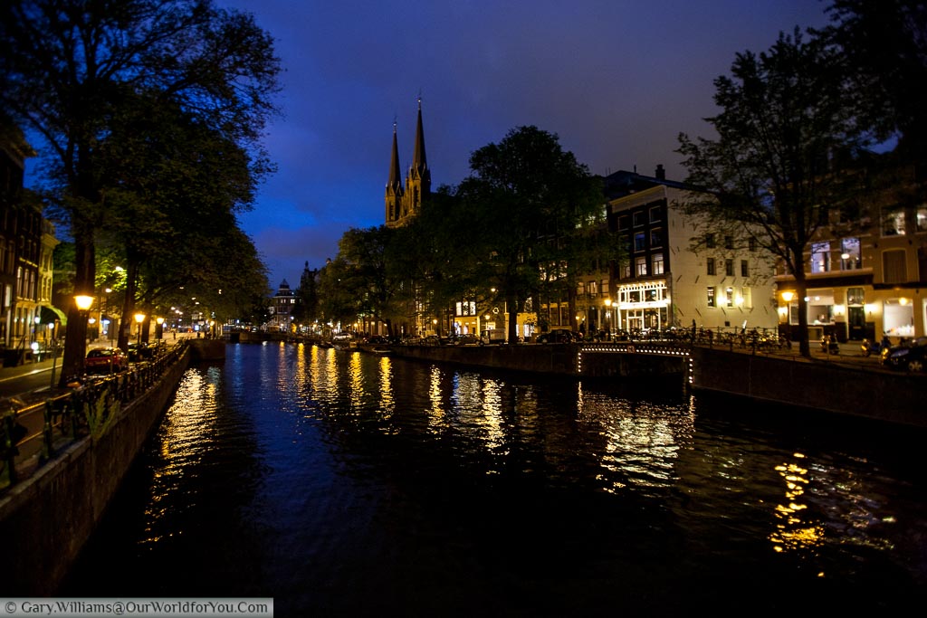 A canal in Amsterdam at dusk with lit streets on either side under a cloudy blue sky.