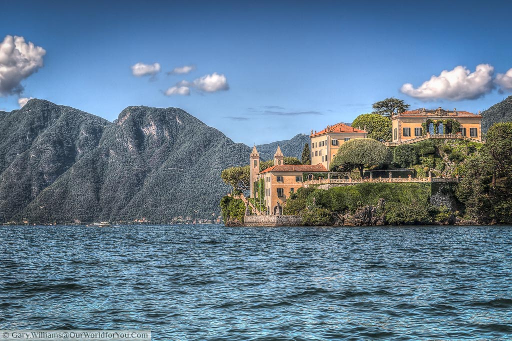 A view from the lake of an ochre coloured Villa with orange tiled roofs set an outcrop on Lake Como with wooded mountains as a backdrop.