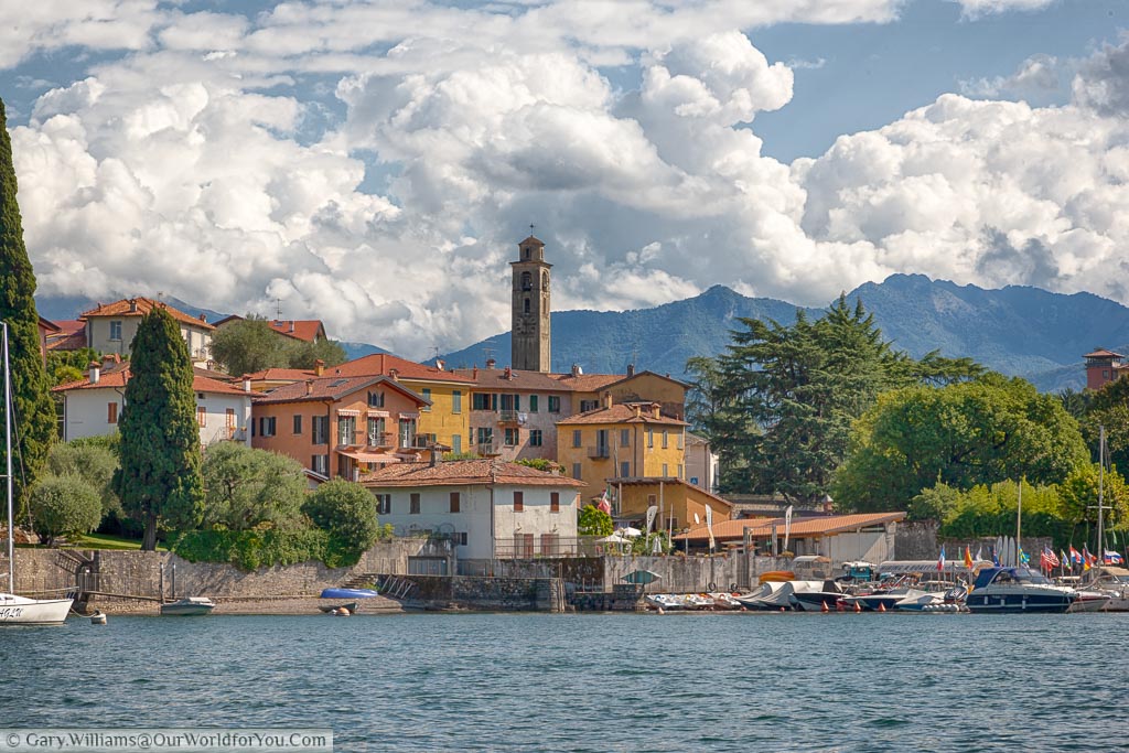 A town on the edge of Lake Como with small harbour at the front and the church tower dominating the skyline against the backdrop of mountains under a lightly cloudy sky.
