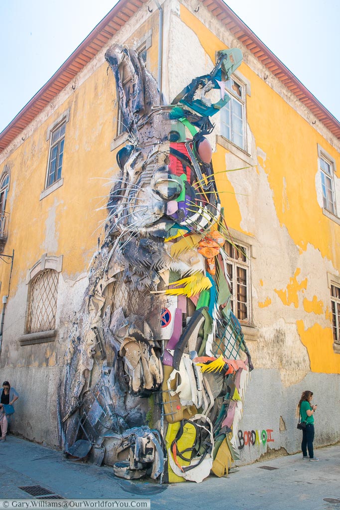 A giant rabbit covering two sides of a corner of a building in the back streets of Porto, Portugal.  The work is constructed of recycled element and reaches to the roofline of the building.