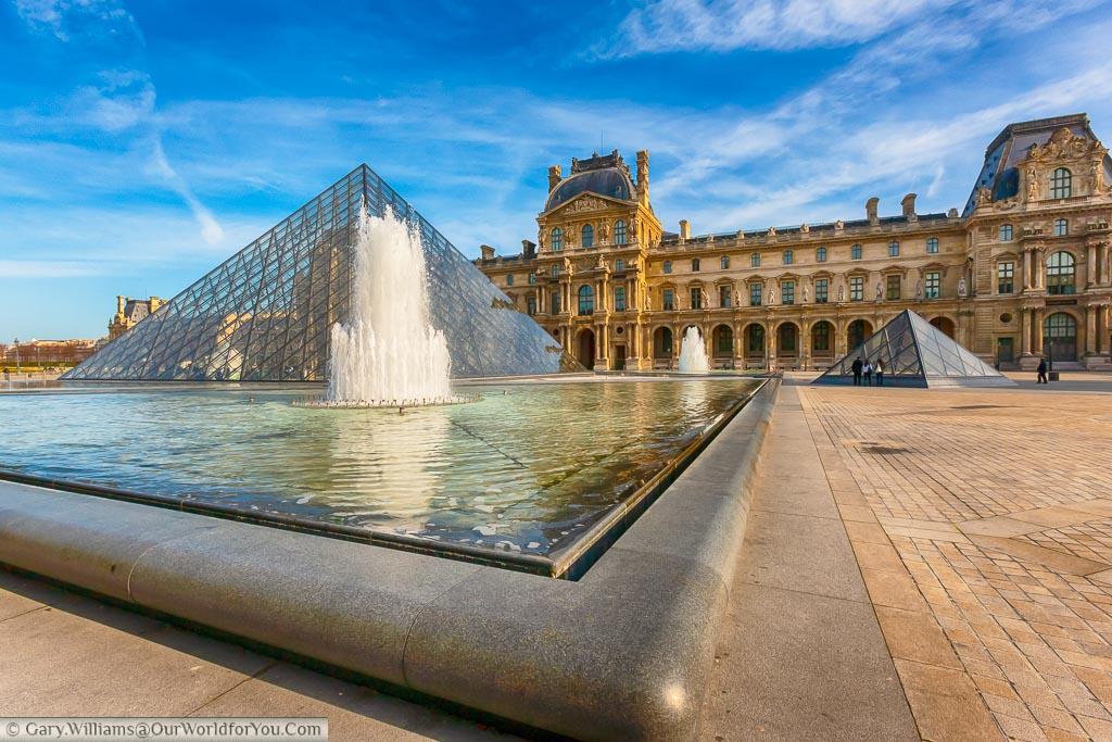 The outside of the Louvre from the corner of the pond with a fountain in front of the pyramid. It's a bright day in Paris, and suprisingly it's quiet in the Napoléon court in front of the museum.