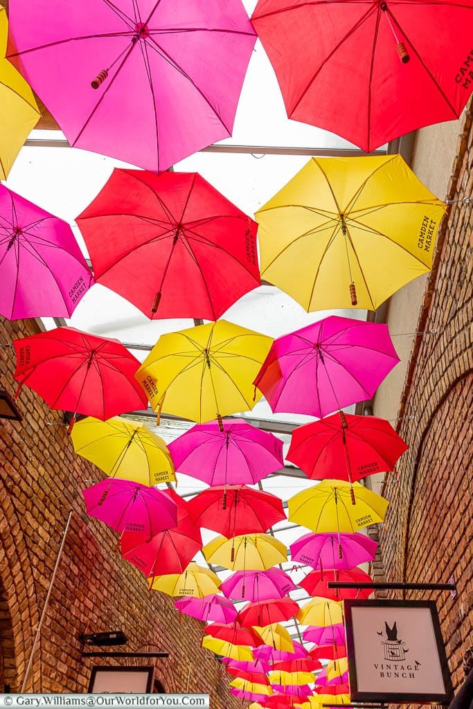 A mixture of pink, yellow & red opened umbrellas provide a canopy between the stores in one lane in Camden Market.