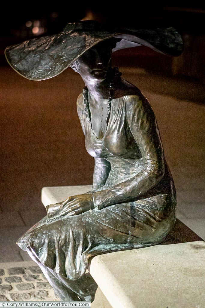 A brass statue of a lady seated on a stone bench with a broad-rimmed hat known as La Dame au Chapeau in Troyes, France