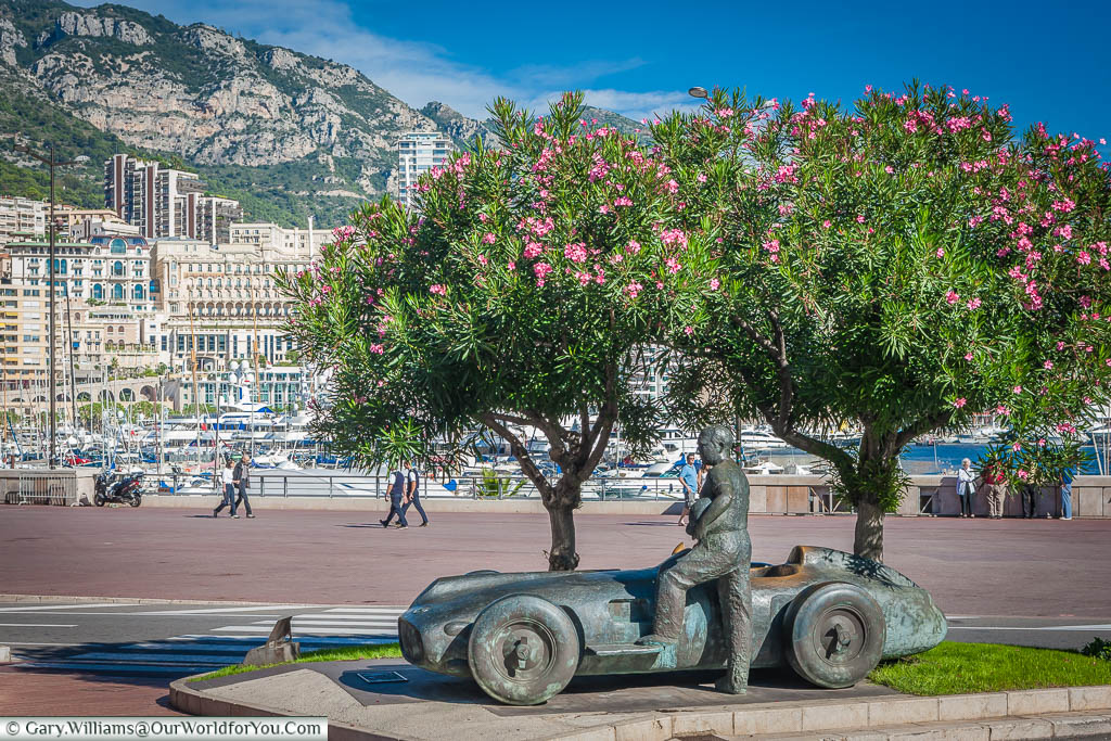 A bronze statue of a facing driver standing next to his 1950's formula 1 car under a couple of trees in flower, in front of the famous Monaco harbour.