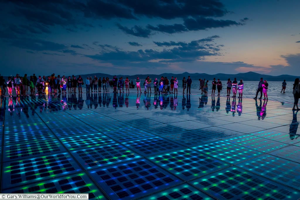People gather around the ‘Greeting to the Sun’ light display at Zadar’s Riva at dusk.  They are illuminated by the LED floor tiles that make up this open-air art installation.