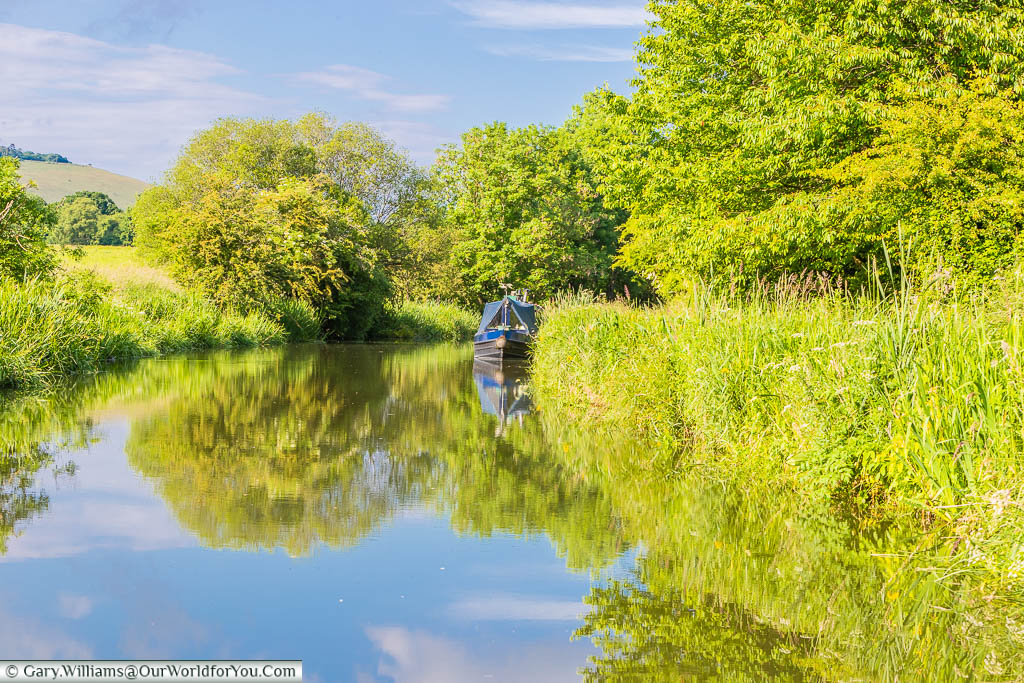 A narrowboat moored in the canalside, in a lush green scene under a blue sky, on the Kennet & Avon Canal.