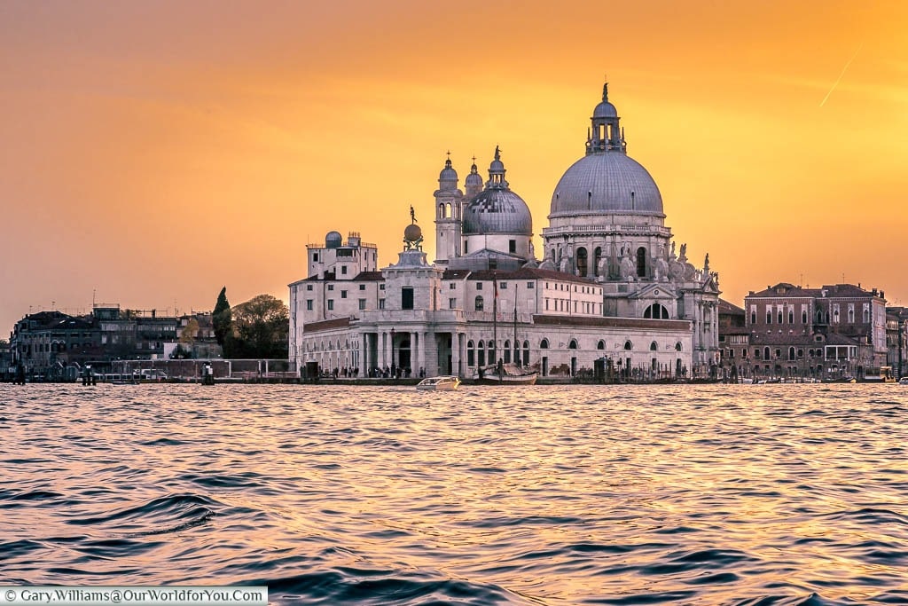 A shot of Santa Maria della Salute at dusk under an orange sky taken from a boat on the lagoon
