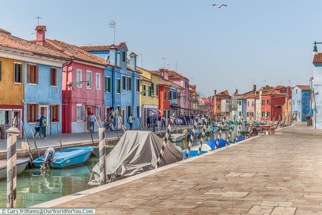 A view along a canal in Burano in which each home is brightly coloured, and small motorboats fill the waterway.