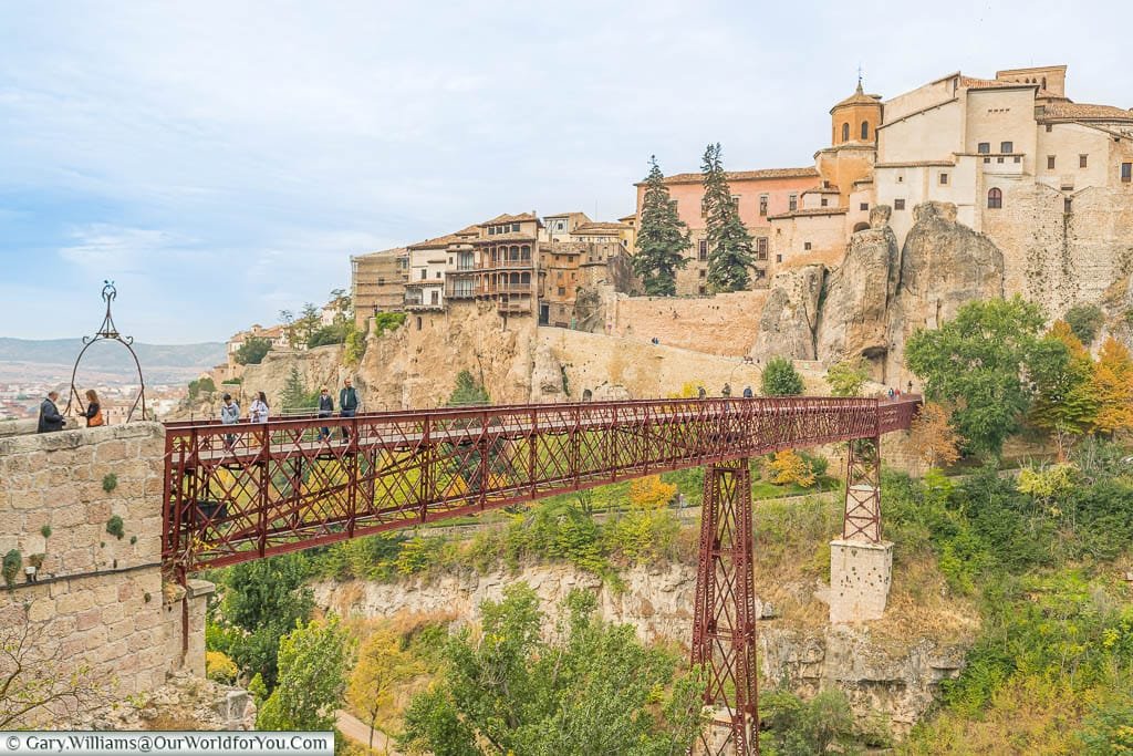 The maroon red wrought iron lattice bridge over a deep ravine to the hillside city of Cuenca with its hanging houses jutting from the rock face.