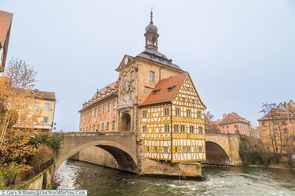 The old town hall of Bamberg, or Altes Rathaus Bamberg. A Baroque style building in the centre of a fast-flowing river connected on either side by two stone arched bridges.
