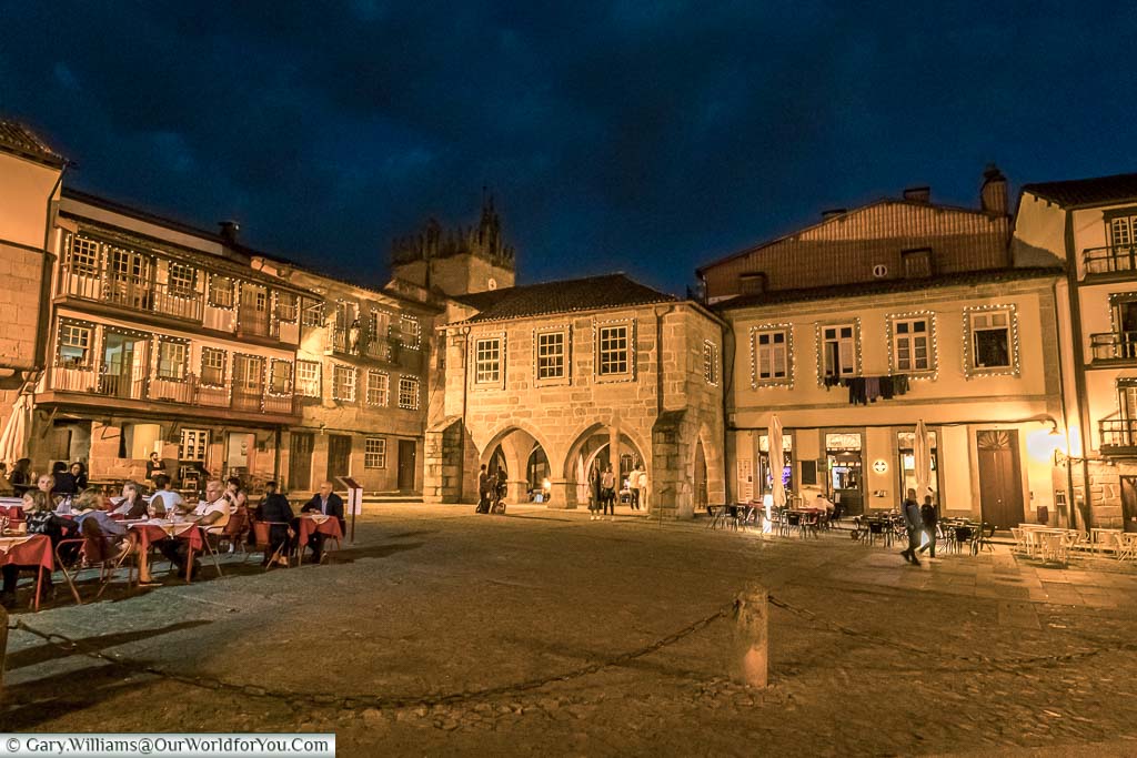 A lit square after dark in Guimarães, Portugal where diners sit at restaurants lining the square and others walkthrough the historical old town.