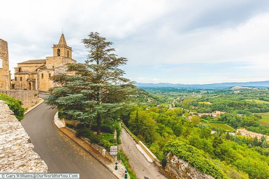 A view of the Provence landscape to the right with a road on the left leading up to the small stone church of Venasque.