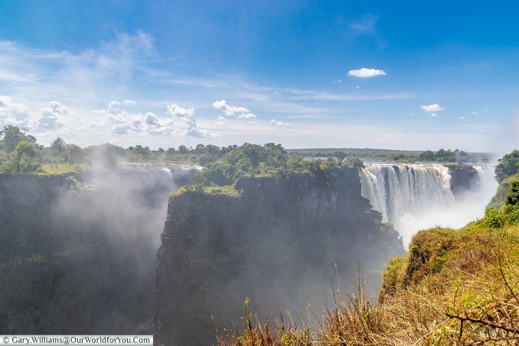 A view of Livingstone Island sits in the flow of the Zambezi between the Victoria Falls. Behind the island, the main falls cascades over the edge underneath the deep blue Sky