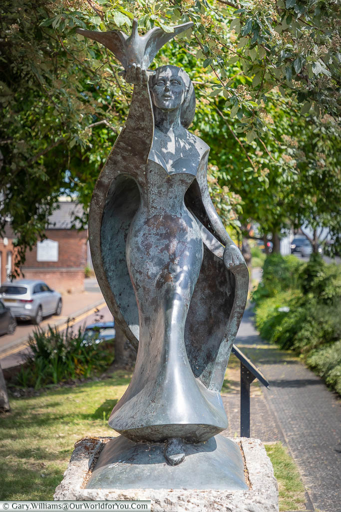 The front view of the modern bronze 'Hope' sculpture depicting a woman stepping forward, releasing a dove of peace, on a green next to West Malling High Street