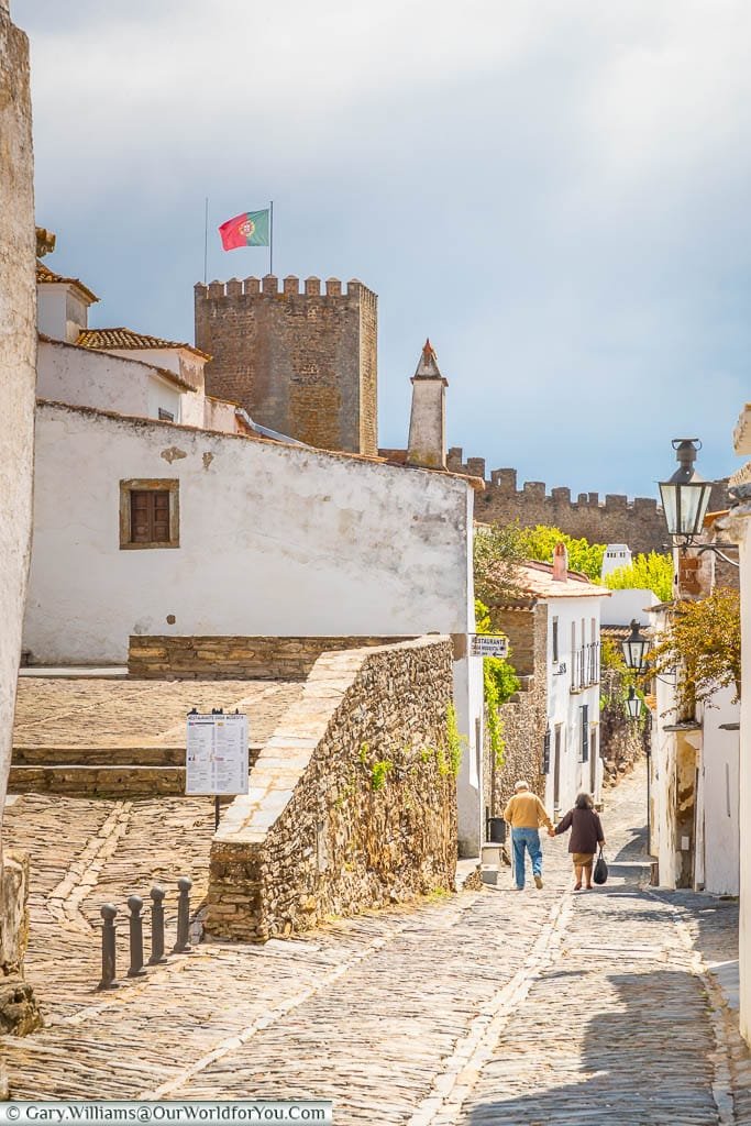 An elderly couple walking along a narrow cobbled lane, with the tower of the town's castle flying the Portuguese flag, in the historic hilltop town of Monsaraz in Portugal